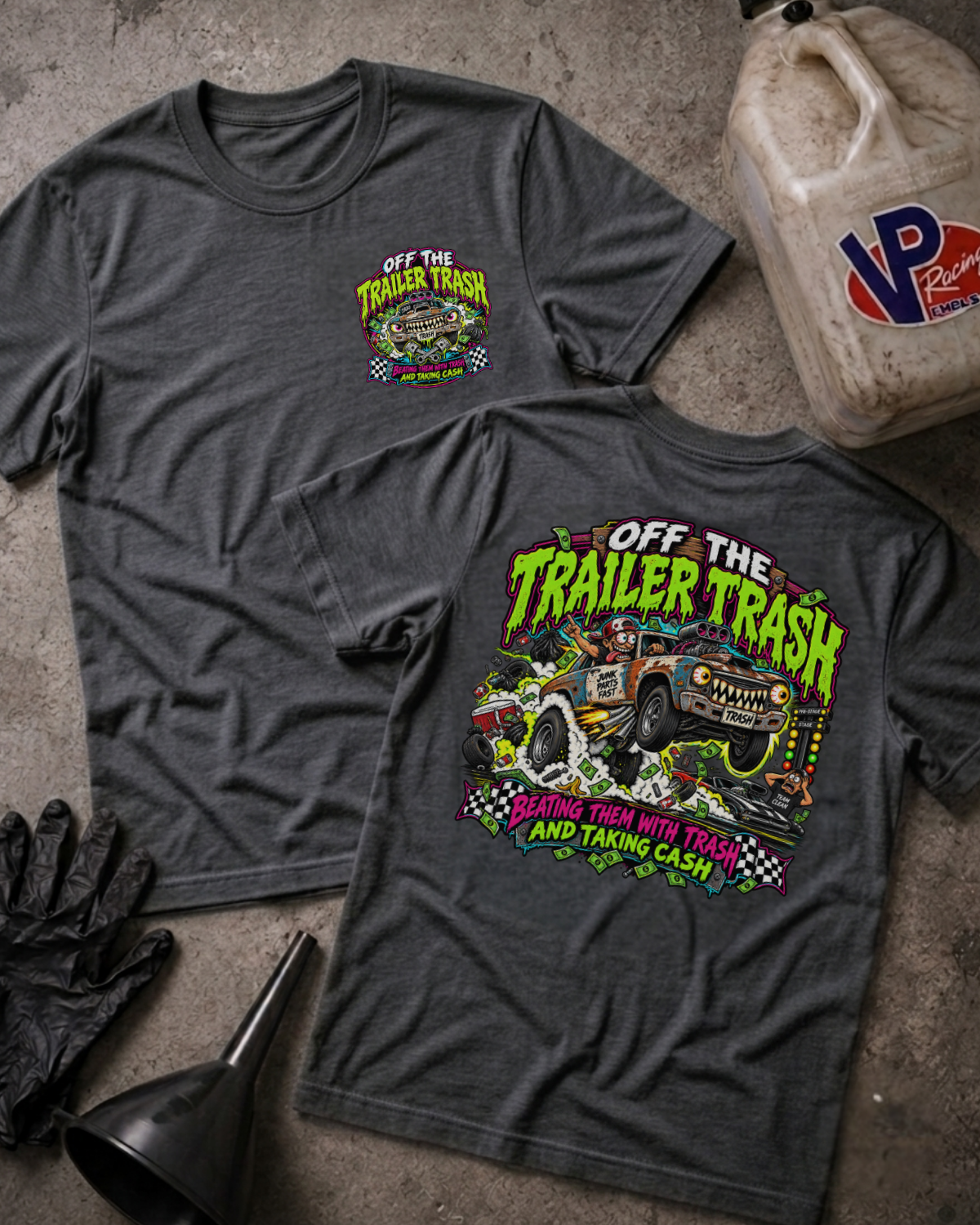 Two dark heather gray graphic T-shirts laid flat on a concrete surface, with a black funnel, black gloves, and a plastic jerrycan nearby. The front shirt shows a small chest graphic reading 'Off The Trailer Trash,' and the back shirt shows a large colorful graphic with the same text and a truck illustration.