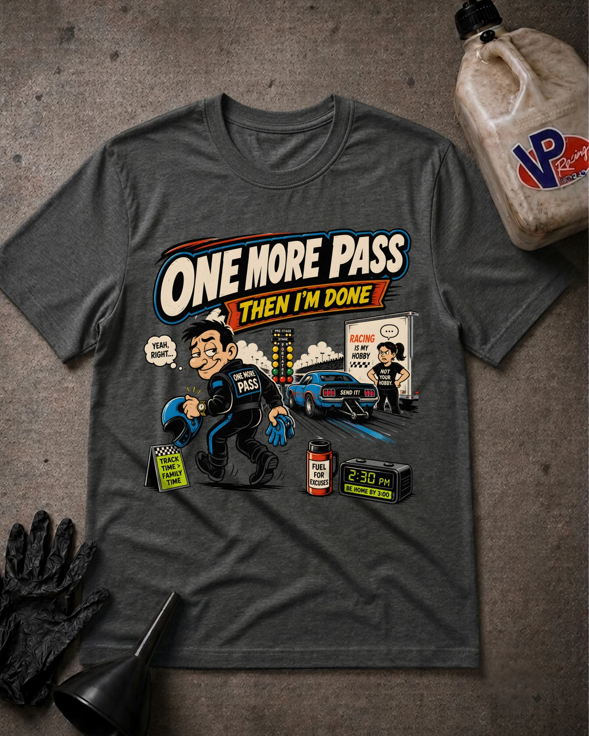 A dark gray graphic t-shirt laid flat on a textured gray surface, surrounded by automotive-themed props including a jug of VP Racing fluid, a black glove, and a funnel. The t-shirt features a colorful cartoon graphic with the text 'ONE MORE PASS THEN I'M DONE' and an illustration of a race car and pit crew.