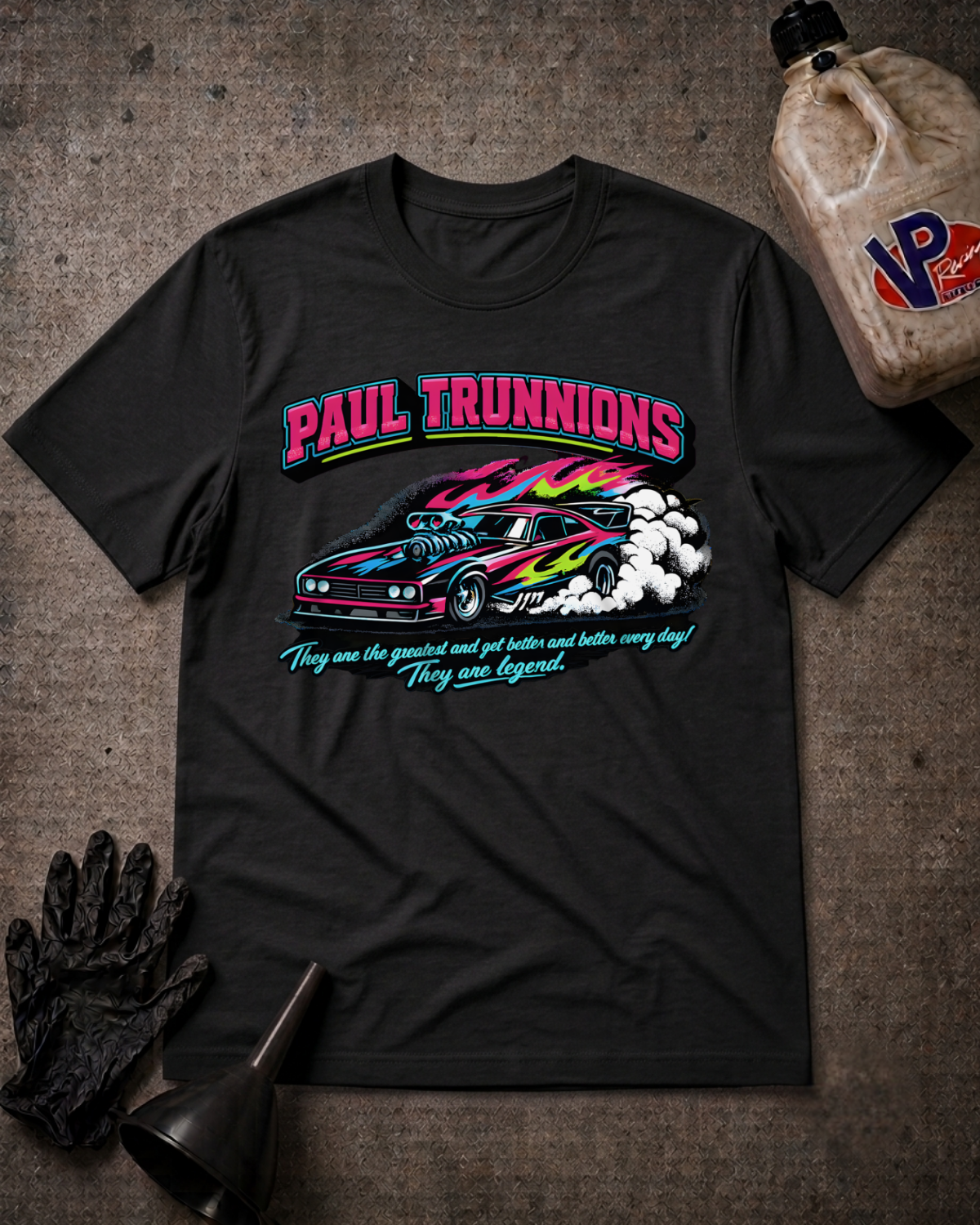 A black graphic t-shirt featuring a colorful print of a race car and the text 'PAUL TRUNNIONS' and 'They are the greatest and get better and better every day! They are legend.' The shirt is displayed in a flat-lay arrangement on a textured gray background with racing-themed props including a bottle of oil, a black glove, and a funnel.
