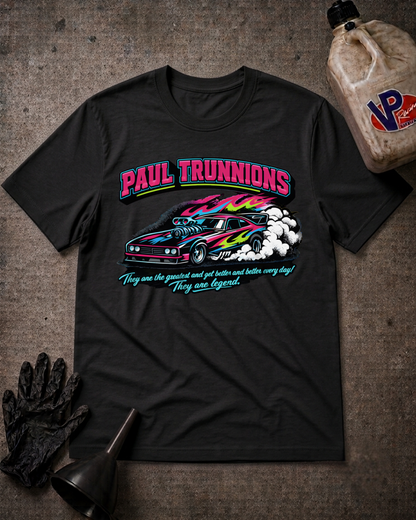 A black graphic t-shirt featuring a colorful print of a race car and the text 'PAUL TRUNNIONS' and 'They are the greatest and get better and better every day! They are legend.' The shirt is displayed in a flat-lay arrangement on a textured gray background with racing-themed props including a bottle of oil, a black glove, and a funnel.