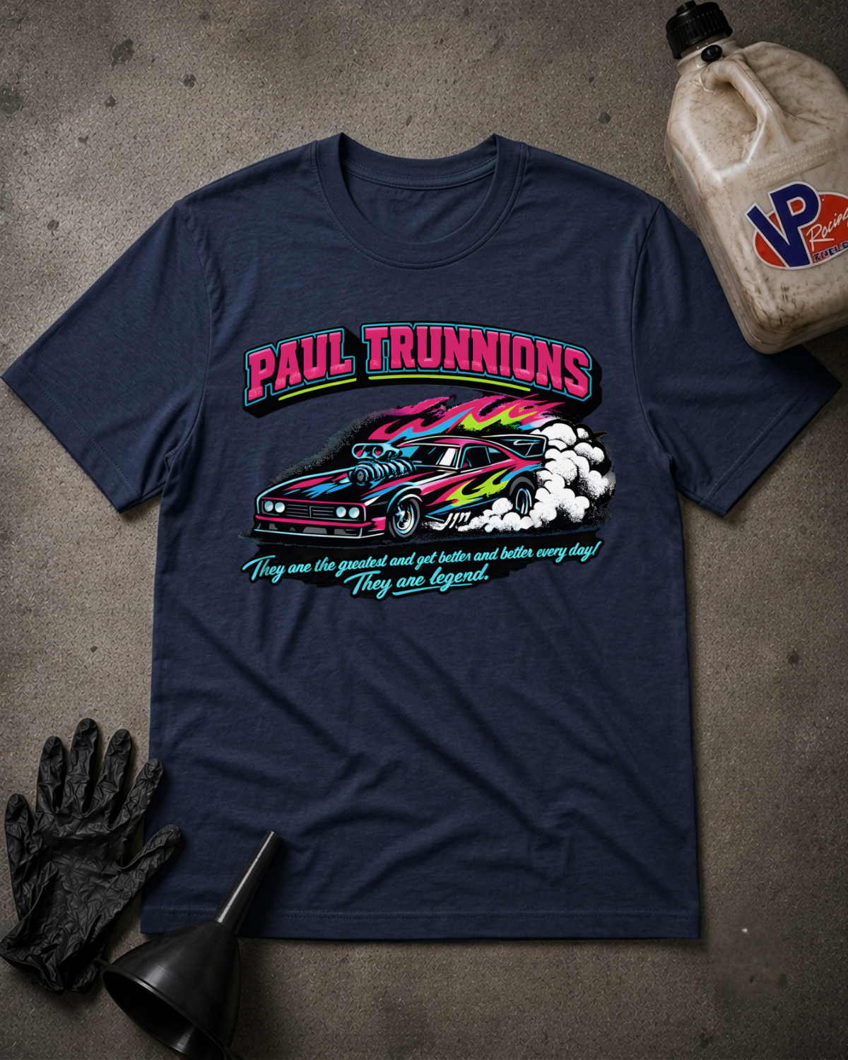 A navy blue graphic t-shirt featuring a Paul Trunnions car design, styled in a flat-lay with a bottle of oil, gloves, and a funnel on a concrete floor.