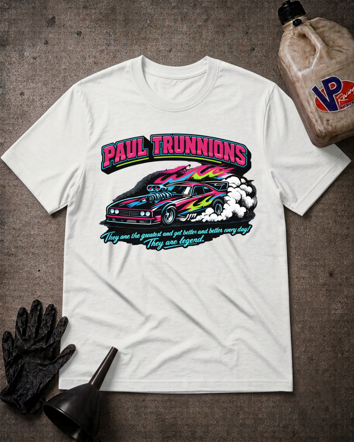 A white graphic t-shirt featuring a colorful 'Paul Trunnions' drag racing design, styled in a flat-lay with black racing gloves, a funnel, and a bottle of oil on a dark textured background.