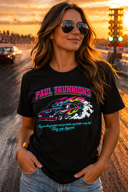 Woman wearing a black t-shirt with colorful graphics and 'Paul Trunnions They are the greatest and get better and better every day! They are lengend' text, standing on a drag strip at sunset.