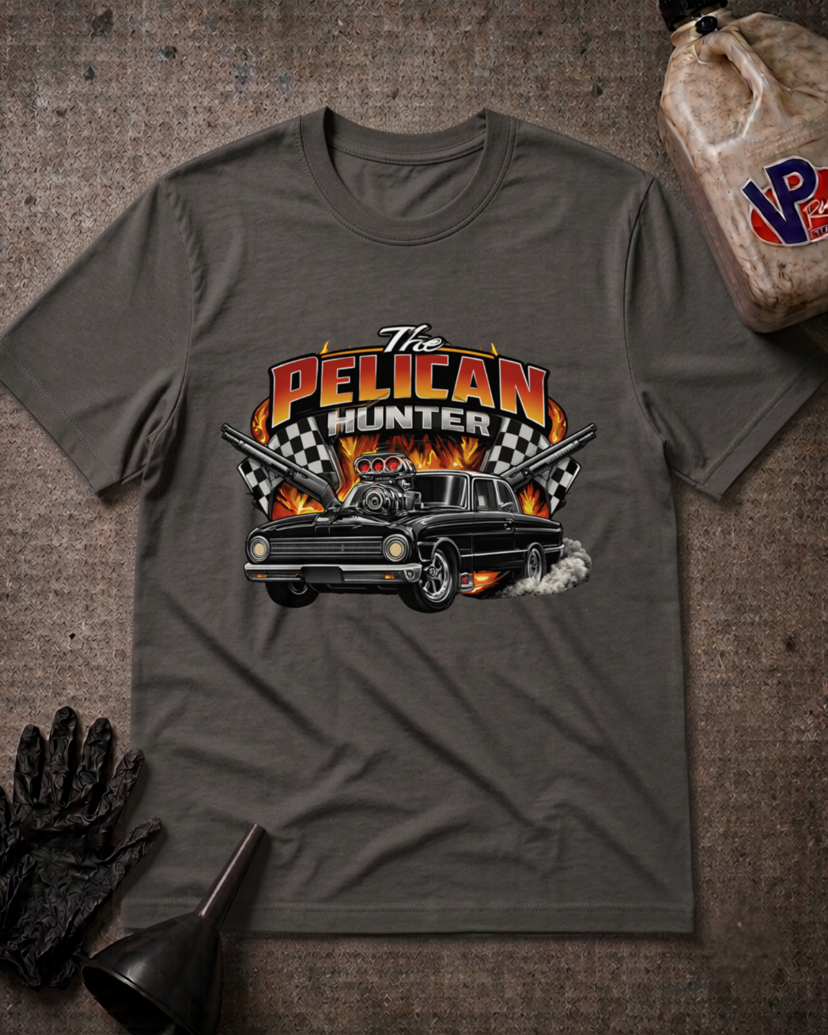 A dark gray graphic t-shirt featuring a 'The Pelican Hunter' design with a black car, flames, and checkered flags, styled flat-lay on a textured gray background with workshop props.
