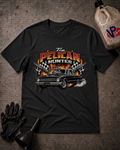 A black graphic t-shirt laid flat on a textured gray background, surrounded by automotive-themed props including an oil bottle, a black funnel, a black glove. The t-shirt features a graphic of a classic car with flames and the text 'The PELICAN HUNTER'.