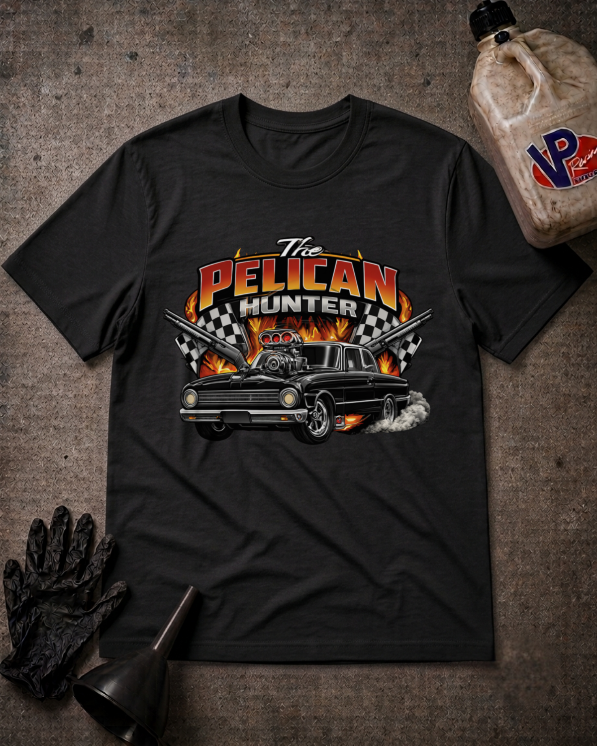 A black graphic t-shirt laid flat on a textured gray background, surrounded by automotive-themed props including an oil bottle, a black funnel, a black glove. The t-shirt features a graphic of a classic car with flames and the text 'The PELICAN HUNTER'.