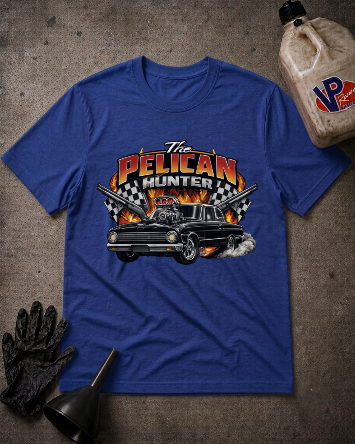 A royal blue graphic t-shirt laid flat on a textured gray surface, surrounded by garage-themed props including a black glove, funnel, and a plastic jug. The t-shirt features a large graphic with the text 'The PELICAN HUNTER' and a black classic car with flames and checkered flags.