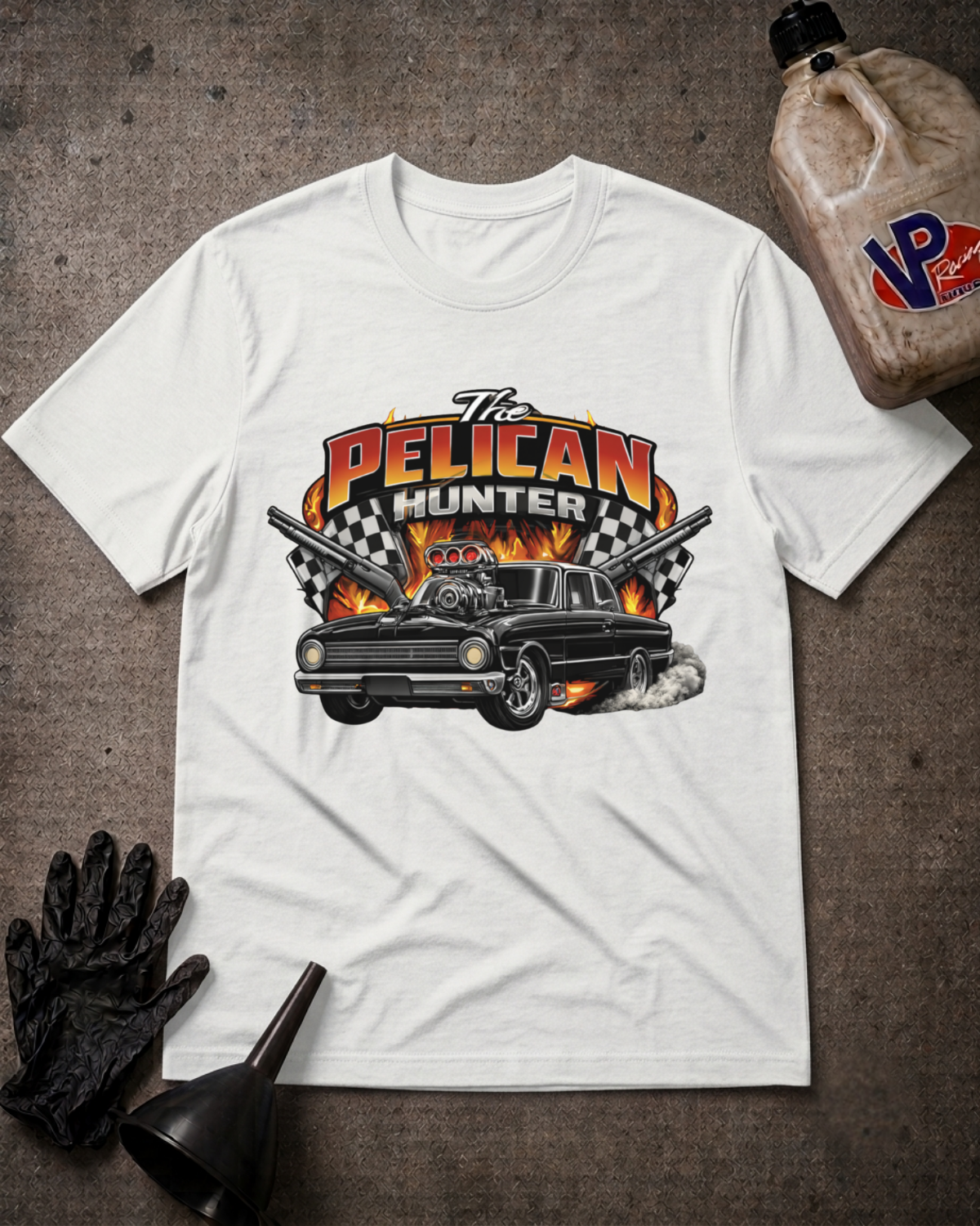A white short-sleeve graphic T-shirt laid flat on a textured brown surface, featuring a large 'The Pelican Hunter' print with a black car, flames, and crossed guns. Black gloves and a black funnel sit beside the shirt, and a plastic jug is partially visible in the top right.