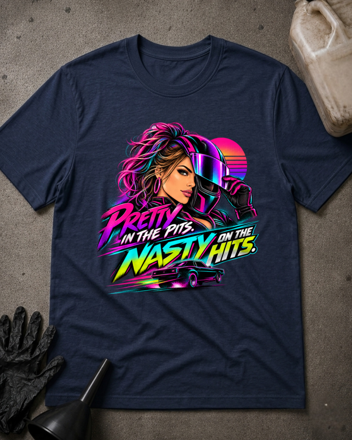 Navy T-shirt laid flat with a neon woman racer, racing helmet, muscle car and sunset graphic reading “Pretty in the Pits. Nasty on the Hits.”