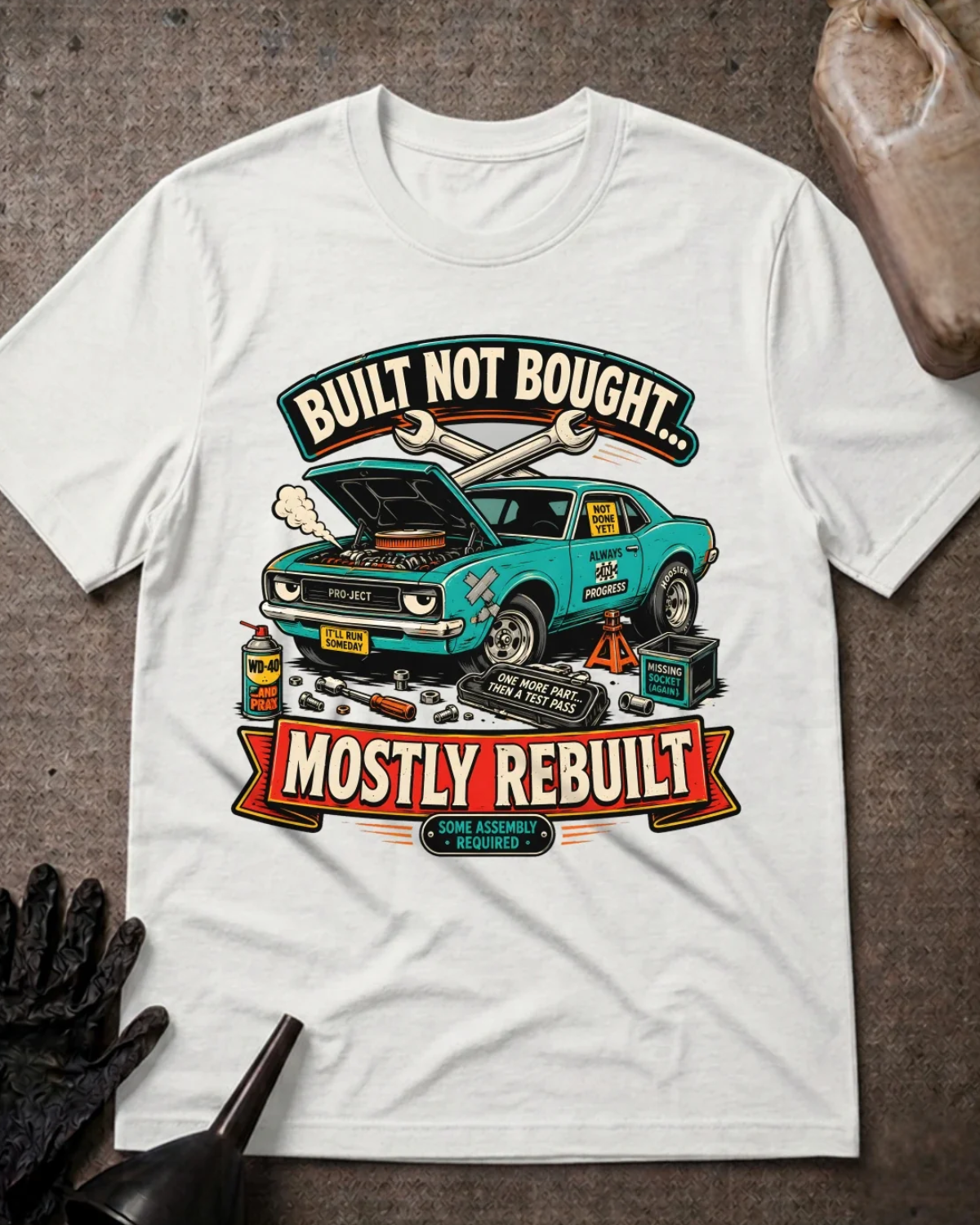 Front of white T-shirt with a teal project car, open hood and tools graphic reading “Built Not Bought… Mostly Rebuilt.”