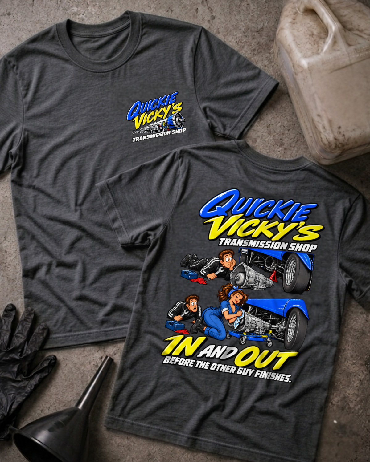 Dark grey heather Quickie Vicky’s Transmission Shop T-shirt showing the front chest logo and back cartoon of a woman mechanic rebuilding a transmission under a blue hot rod, “In and Out Before the Other Guy Finishes,” with gloves, funnel, and oil jug.