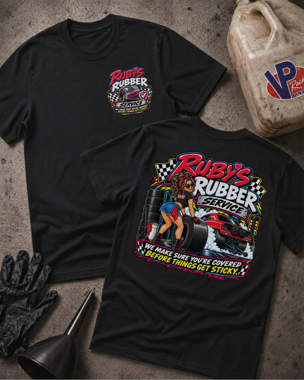 Two black graphic T-shirts laid flat on a concrete floor, showing the front and back designs for 'Ruby's Rubber Service'. The front features a small chest logo, while the back displays a large graphic with a woman, tires, and a race car.