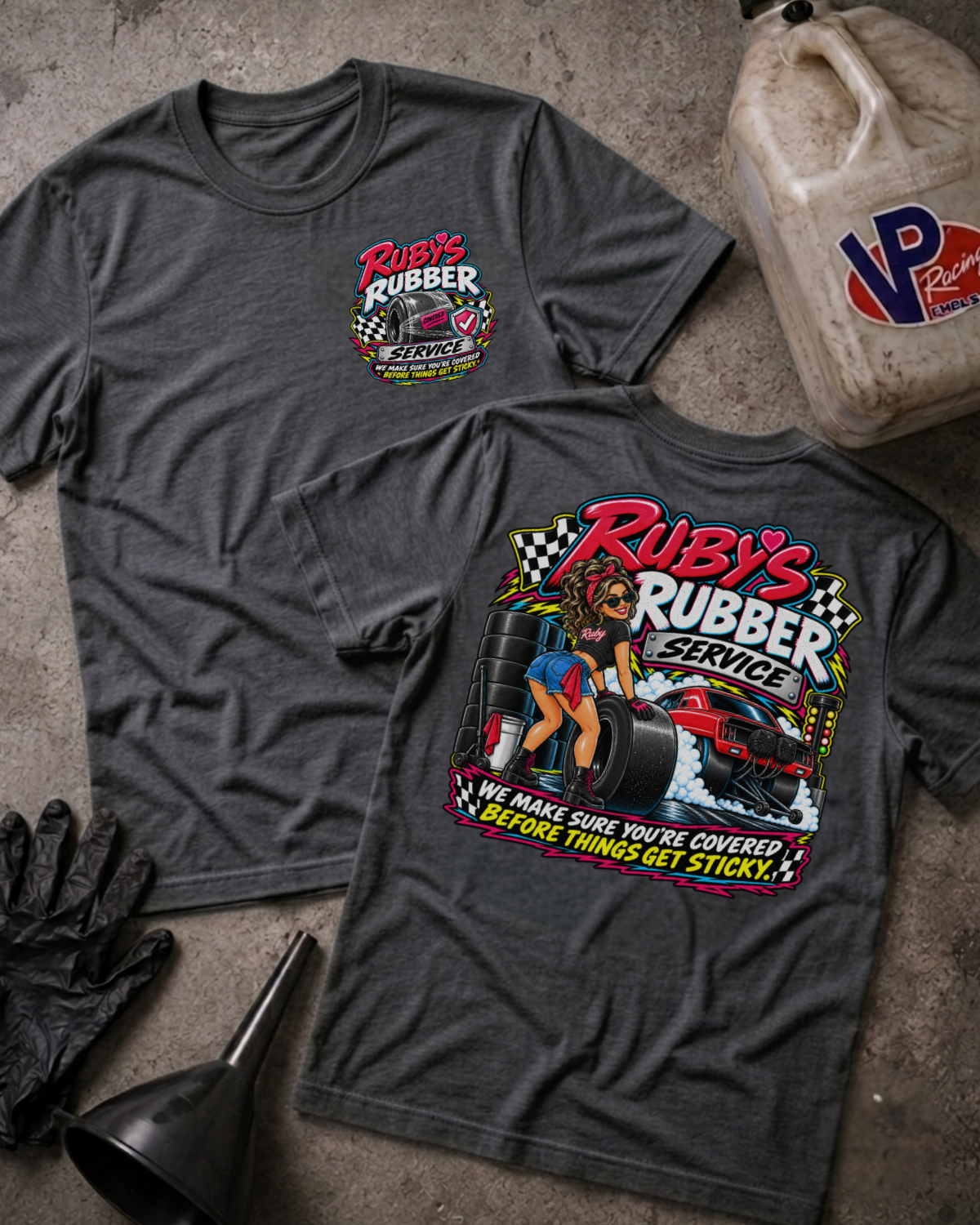 Two dark heather gray graphic T-shirts laid flat on a concrete-like surface, with a vintage oil can, black gloves, and a funnel around them. The front shirt shows a large colorful 'Rubby's Rubber Service' racing graphic, and the back shirt shows a smaller matching chest graphic.