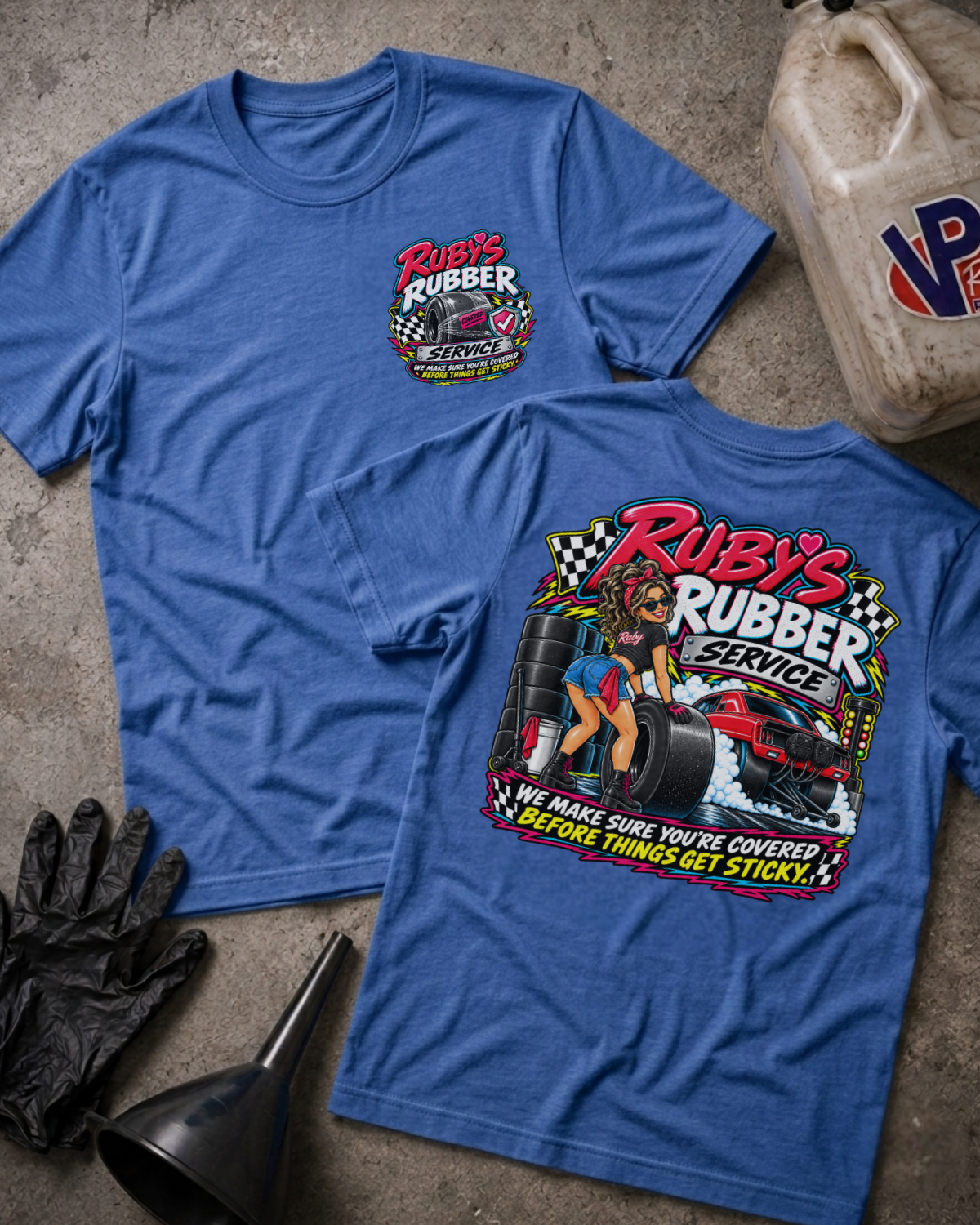 Two heather blue graphic T-shirts laid flat on a concrete floor, showing the front and back designs for 'Ruby's Rubber Service'. The front features a small chest logo, while the back displays a large graphic with a woman, tires, and a race car.