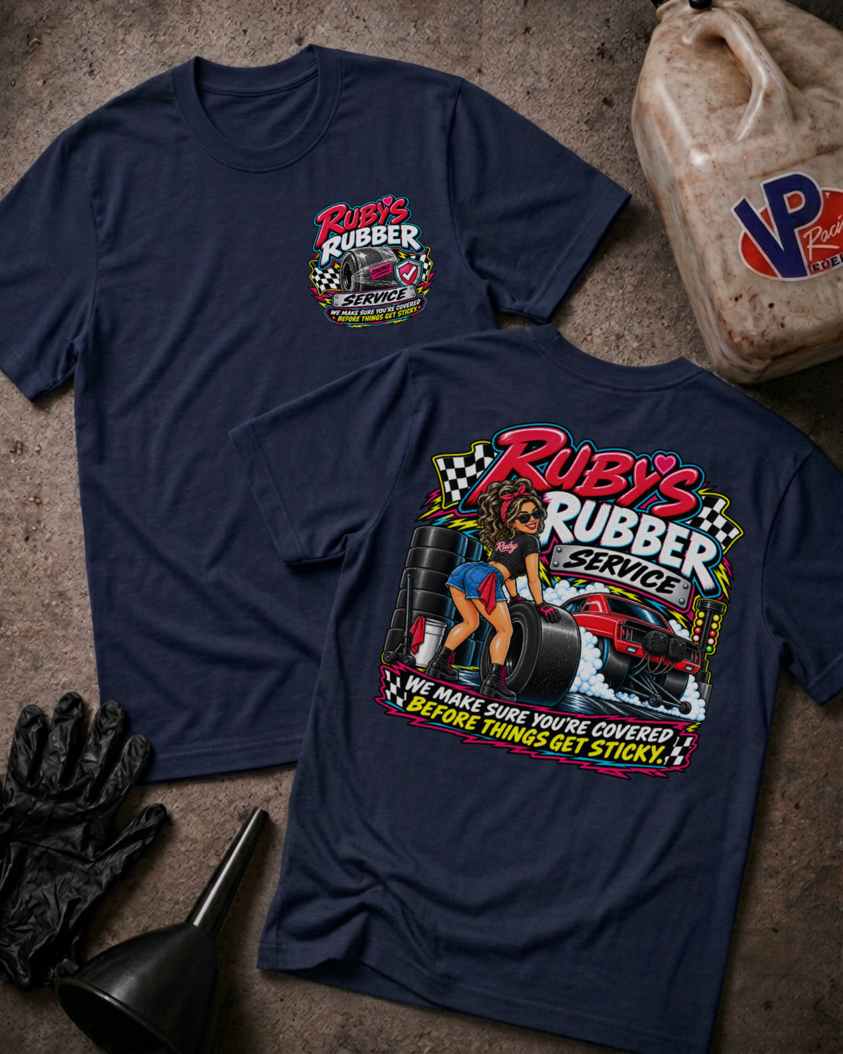 Two navy graphic T-shirts laid flat on a concrete floor, showing the front and back designs for 'Ruby's Rubber Service'. The front features a small chest logo, while the back displays a large graphic with a woman, tires, and a race car.