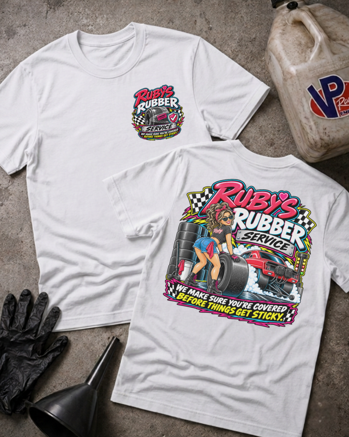 Two white graphic T-shirts laid flat on a concrete floor, showing the front and back designs for 'Ruby's Rubber Service'. The front features a small chest logo, while the back displays a large graphic with a woman, tires, and a race car.