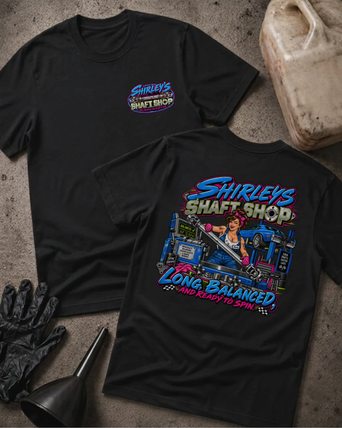 Front and back of Shirley’s Shaft Shop black T-shirt with chest logo, woman holding a driveshaft in a workshop, and “Long, balanced, and ready to spin” slogan.