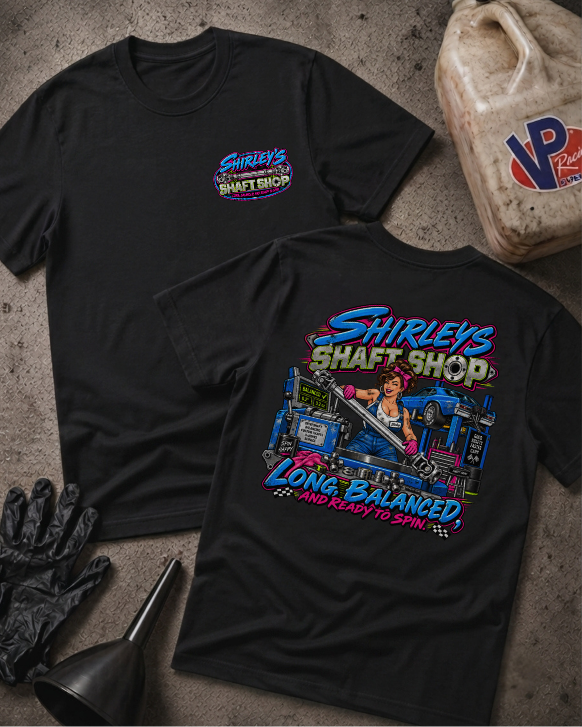 Two black graphic t-shirts with a 'SHIRLEY'S SHAFT SHOP' logo on the front and color graphic of a pinup style woman holding a car shaft in a garage with text 'SHIRLEY'S SHAFT SHOP LONG, BALANCED AND READY TO SPIN' on the back