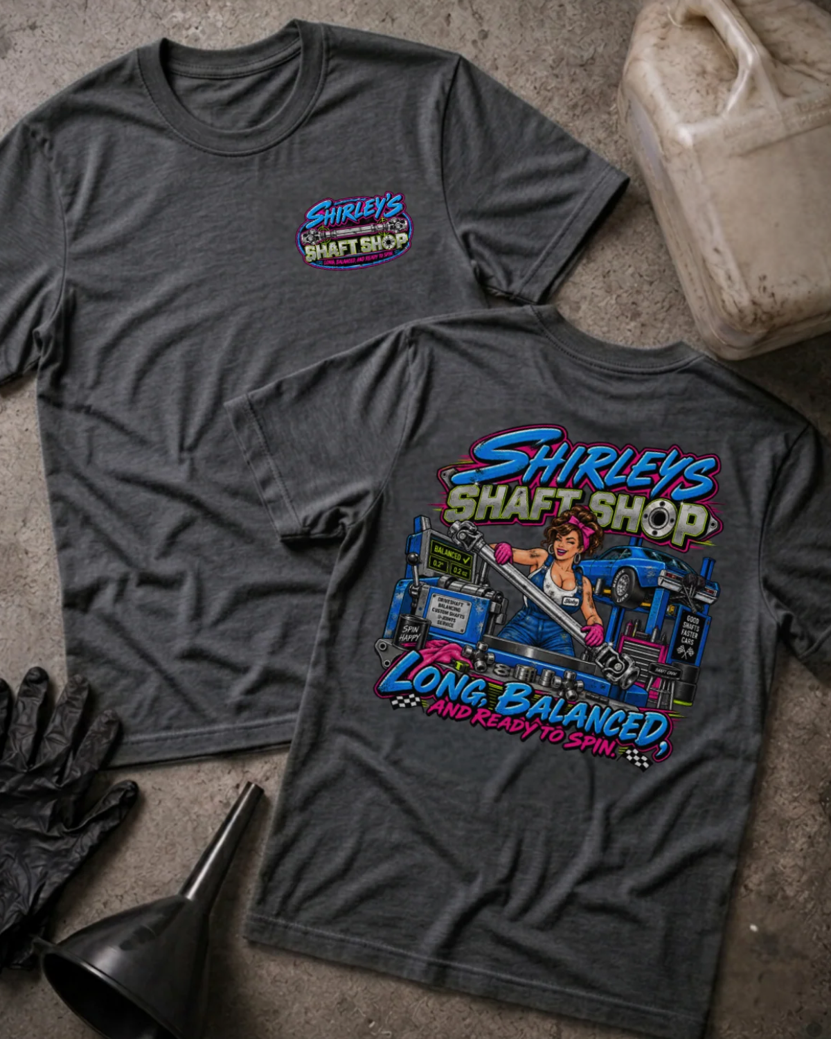 Front and back of Shirley’s Shaft Shop dark grey heather T-shirt with chest logo, woman holding a driveshaft in a workshop, and “Long, balanced, and ready to spin” slogan.