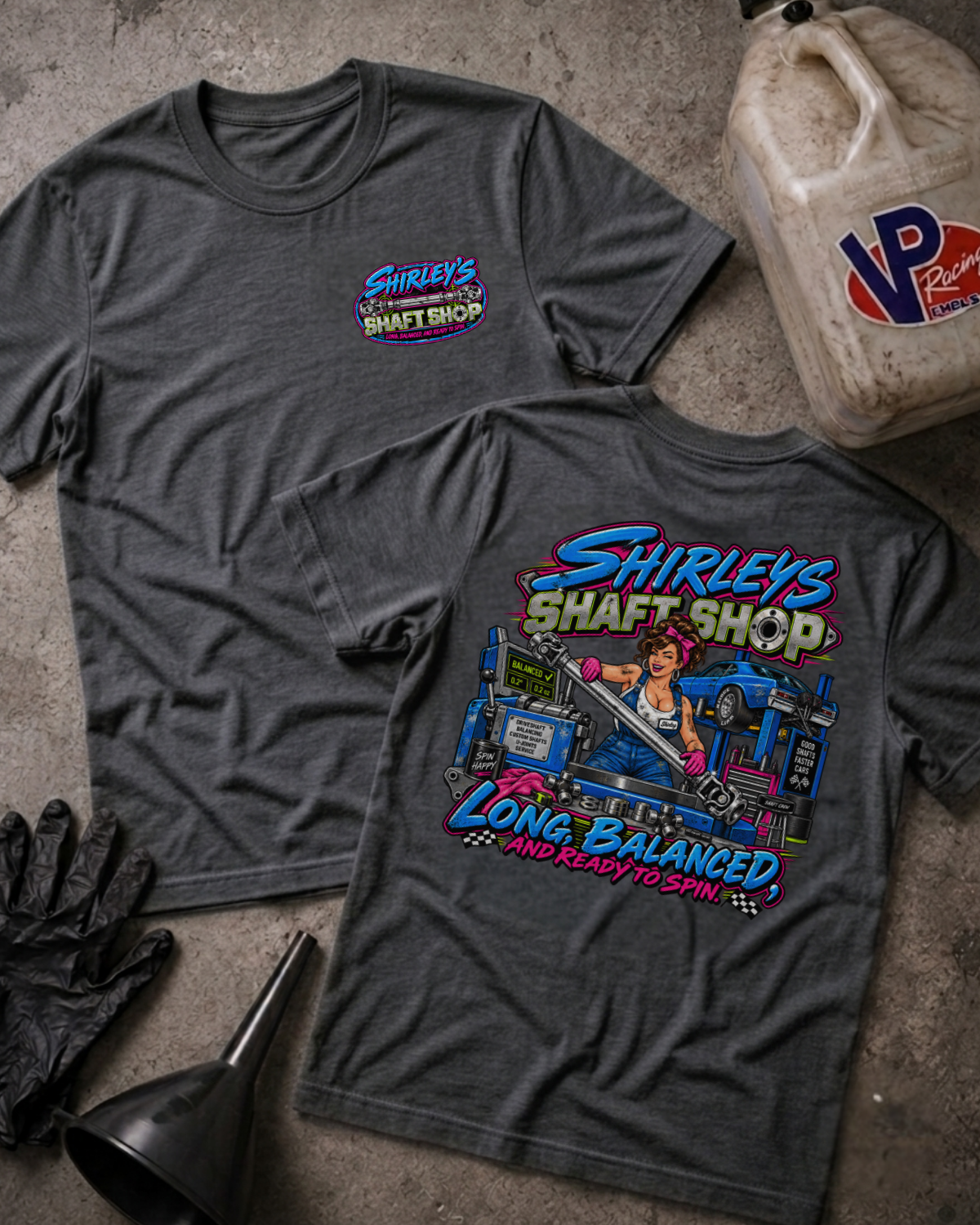Two dark grey heather graphic t-shirts with a 'SHIRLEY'S SHAFT SHOP' logo on the front and color graphic of a pinup style woman holding a car shaft in a garage with text 'SHIRLEY'S SHAFT SHOP LONG, BALANCED AND READY TO SPIN' on the back