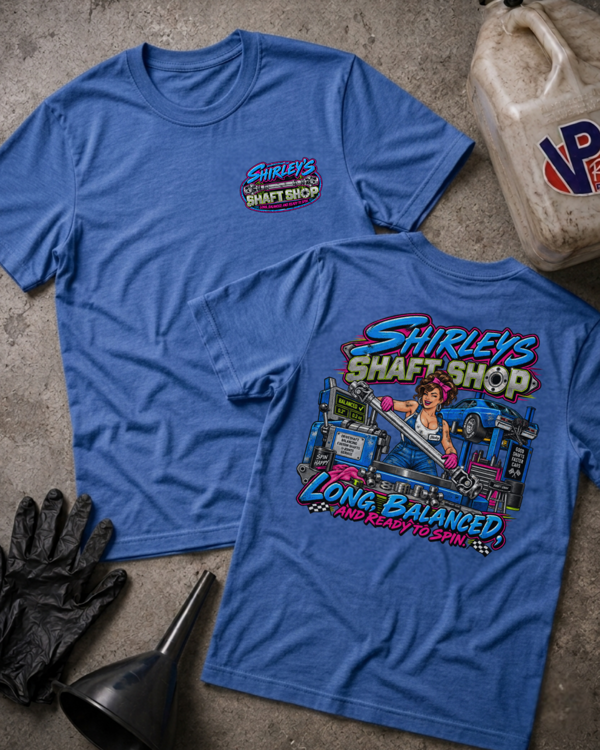 Two heather true royal graphic t-shirts with a 'SHIRLEY'S SHAFT SHOP' logo on the front and color graphic of a pinup style woman holding a car shaft in a garage with text 'SHIRLEY'S SHAFT SHOP LONG, BALANCED AND READY TO SPIN' on the back