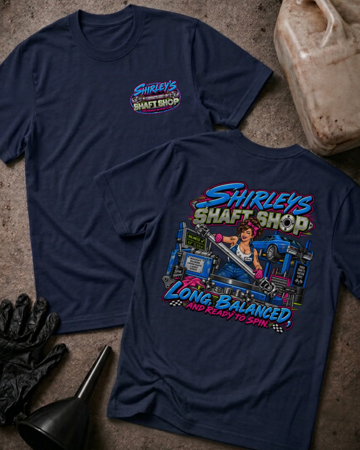 Front and back of Shirley’s Shaft Shop navy T-shirt with chest logo, woman holding a driveshaft in a workshop, and “Long, balanced, and ready to spin” slogan.