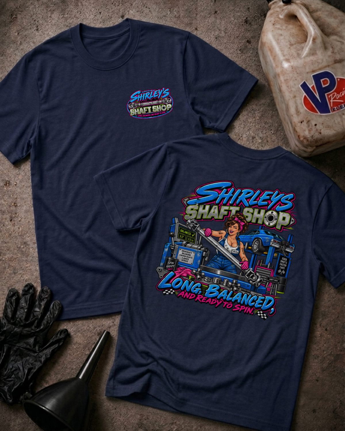 Two navy graphic t-shirts with a 'SHIRLEY'S SHAFT SHOP' logo on the front and color graphic of a pinup style woman holding a car shaft in a garage with text 'SHIRLEY'S SHAFT SHOP LONG, BALANCED AND READY TO SPIN' on the back