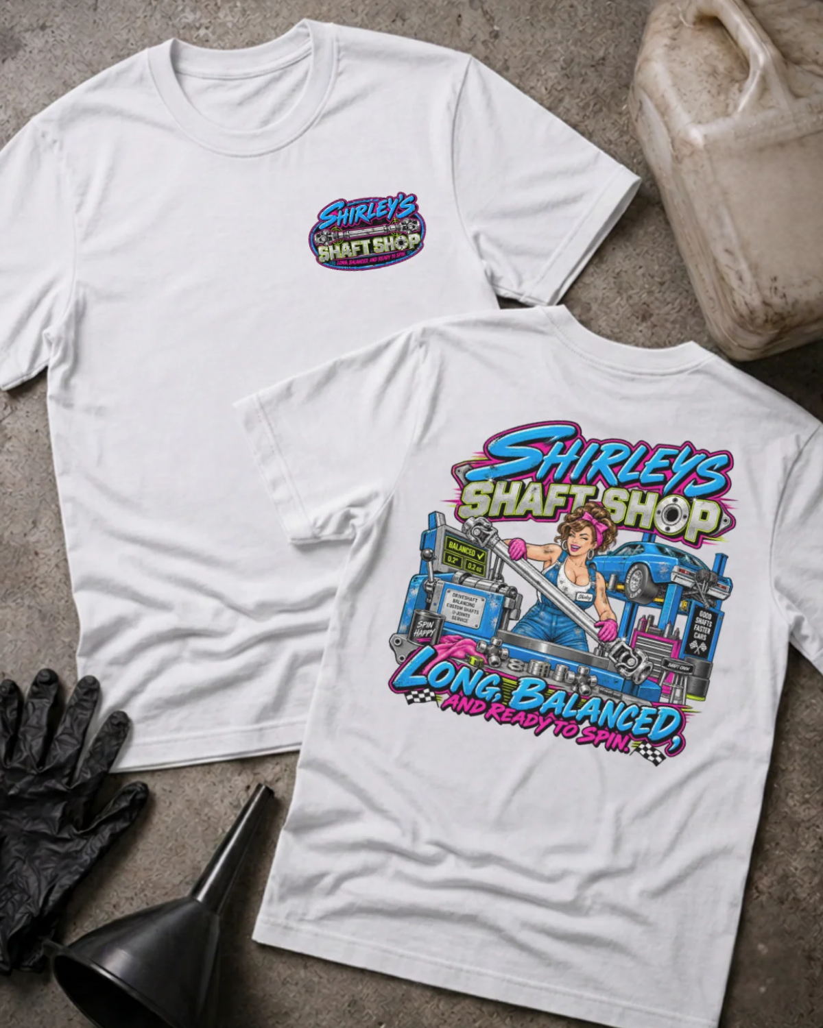 Front and back of Shirley’s Shaft Shop white T-shirt with chest logo, woman holding a driveshaft in a workshop, and “Long, balanced, and ready to spin” slogan.