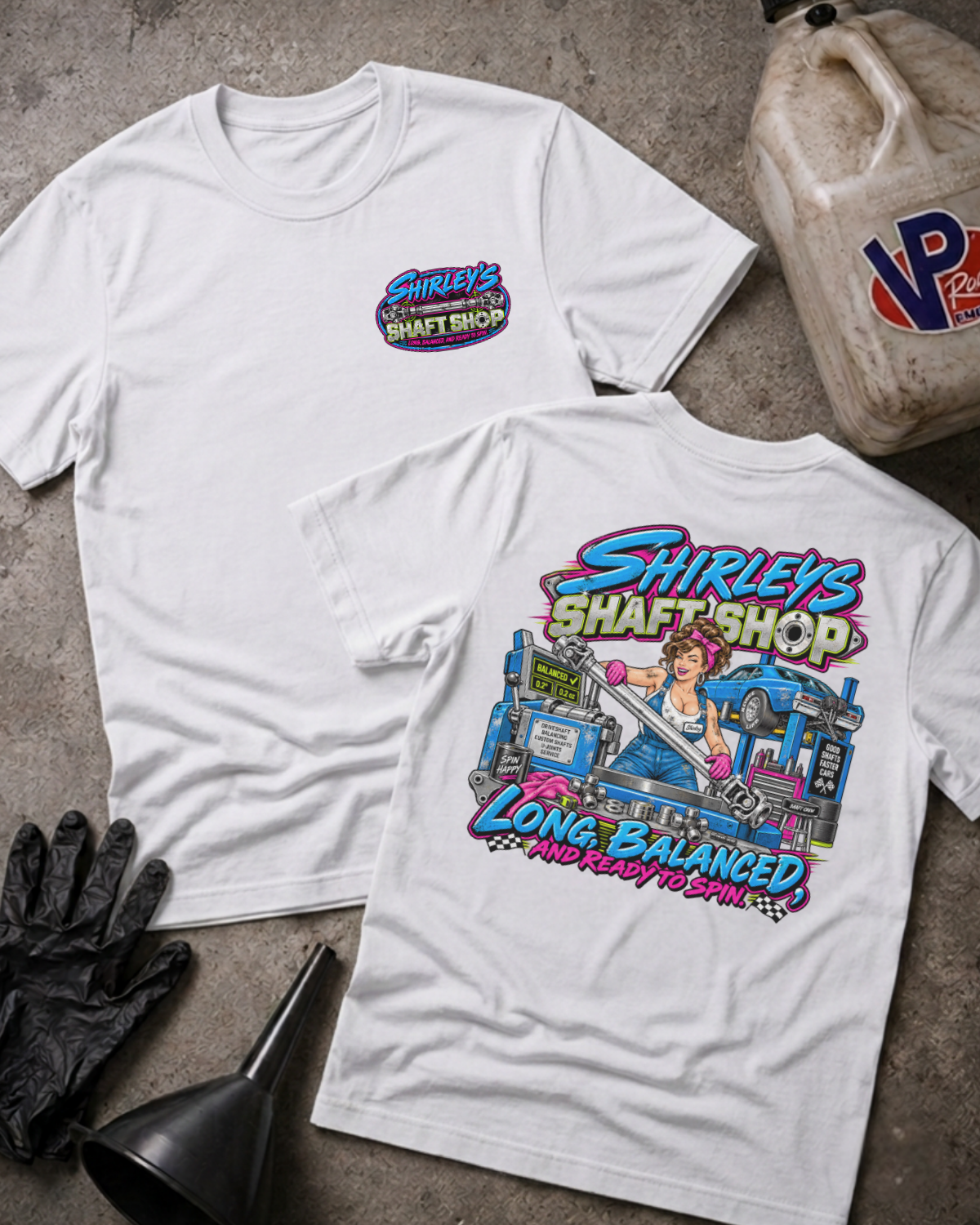 Two white graphic t-shirts with a 'SHIRLEY'S SHAFT SHOP' logo on the front and color graphic of a pinup style woman holding a car shaft in a garage with text 'SHIRLEY'S SHAFT SHOP LONG, BALANCED AND READY TO SPIN' on the back