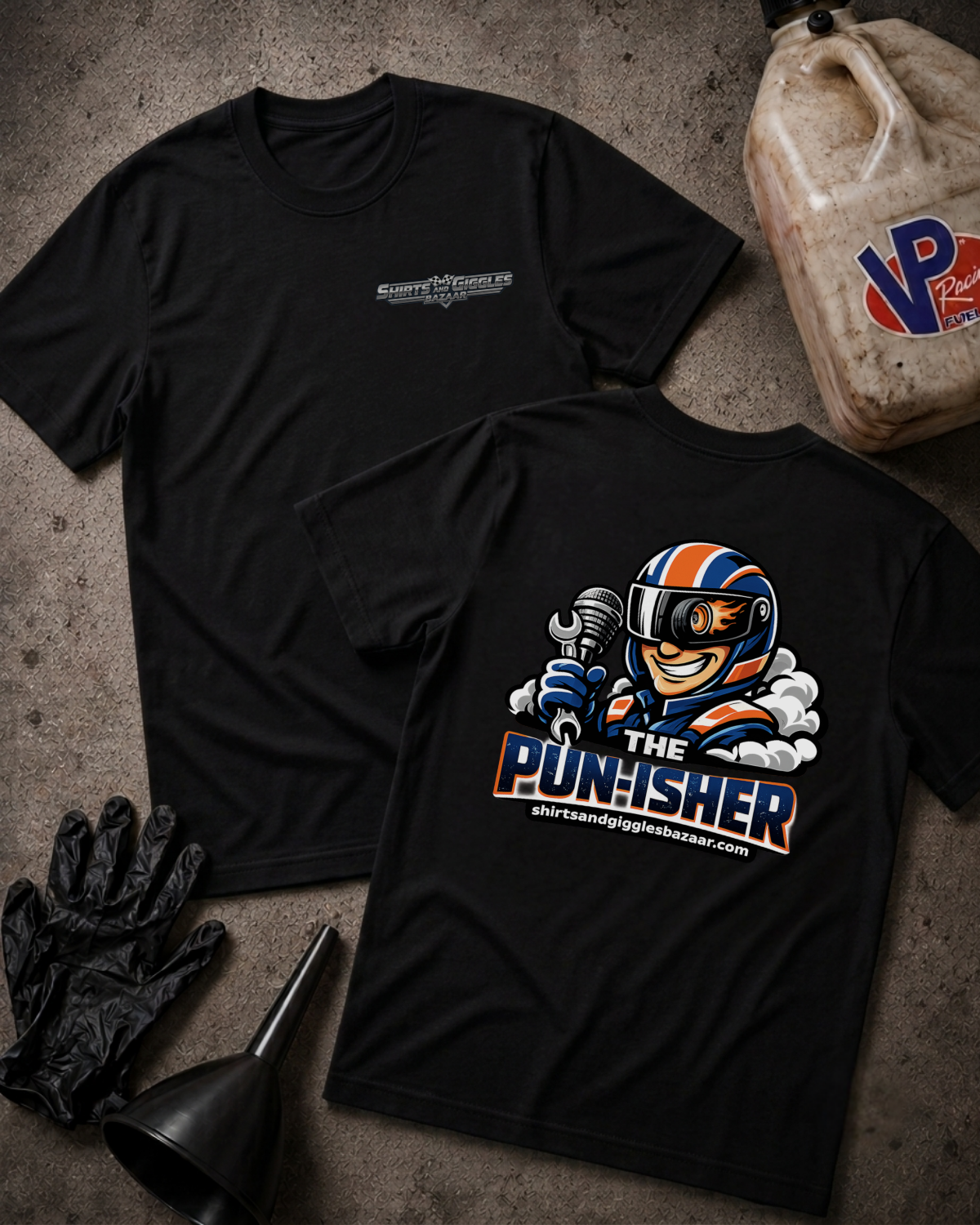 Two black short-sleeve t-shirts displayed on a textured gray surface, with one showing a small chrome chest logo and the other showing a large back graphic of a cartoon race car driver holding a wrench with the text 'THE PUNISHER' and 'shirtsandgigglesbazaar.com'