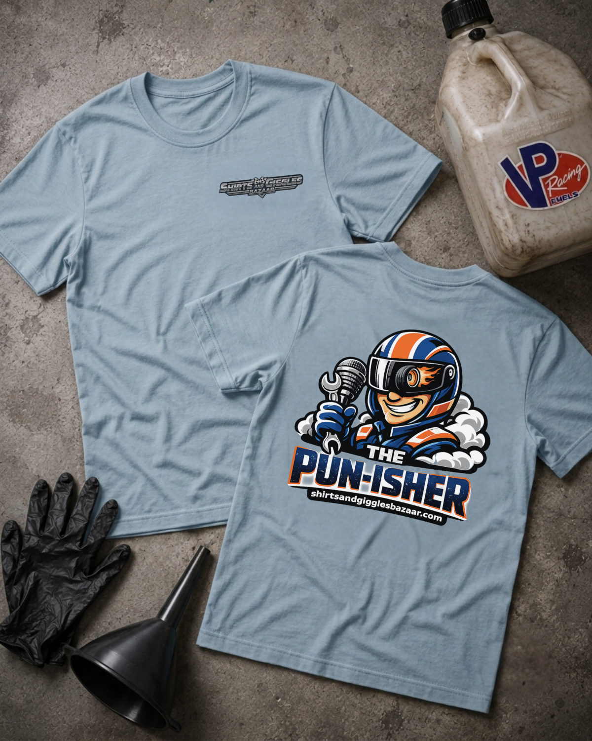 Two light blue graphic T-shirts laid flat on a textured gray surface, with a large cartoon racecar driver graphic on the back and a smaller chest logo on the front. Black gloves, a black funnel, and a VP Racing jug are placed around them as styling props.
