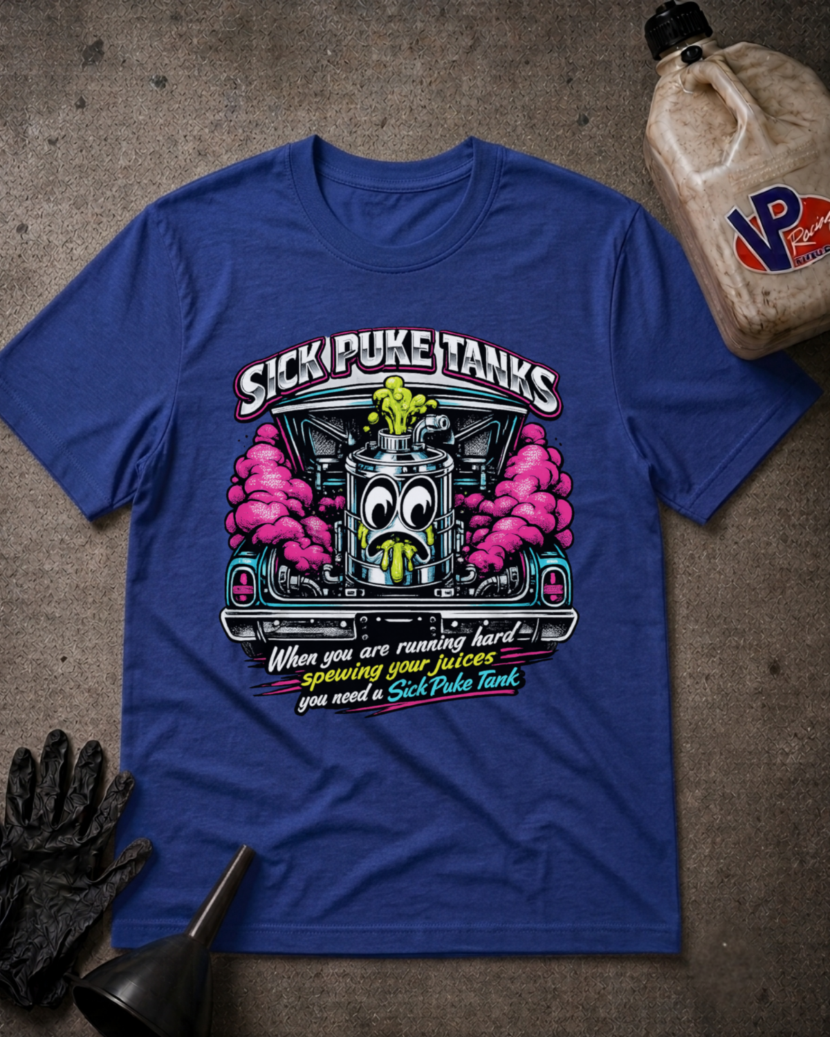 A royal blue graphic t-shirt featuring a cartoon fuel tank with a surprised face and pink smoke, with the text 'SICK PUKE TANKS' above it. The shirt is displayed in a flat-lay arrangement on a textured gray concrete surface, accompanied by a black work glove, a funnel, and a plastic jug of oil.
