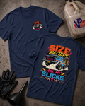 Two navy blue graphic T-shirts laid flat on a textured concrete surface, with a race gas jug, black gloves, and funnel as props. The front shirt shows a small multicolor chest graphic, and the back shirt shows a large colorful racing-themed print with a blue muscle car and the text 'SIZE MATTERS ASK MY SLICKS'.