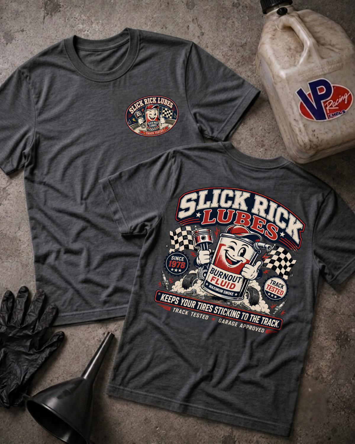 Two dark charcoal gray graphic T-shirts laid flat on a textured concrete surface, showing both front and back designs for Slick Rick Lubes. A beige VP Racing fuel jug, black gloves, and a black funnel are placed around them as styled props.