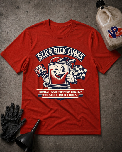 A red graphic t-shirt laid flat on a textured gray surface, surrounded by black mechanic gloves, a black funnel, and a plastic jug of oil. The shirt features a retro-style graphic with the text 'SLICK RICK LUBES' and an illustration of a smiling red lubricant can holding a checkered flag.
