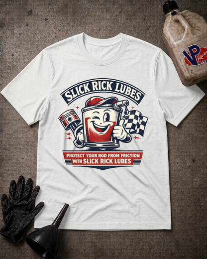 A white graphic t-shirt laid flat on a textured dark gray surface, surrounded by racing-themed props including a black oil bottle, a black funnel, and a black glove. The shirt features a retro-style cartoon mascot of a red and white lubricant can with a smiling face, alongside a checkered flag and the text 'SLICK RICK LUBES' and 'PROTECT YOUR ROD FROM FRICTION WITH SLICK RICK LUBES'.