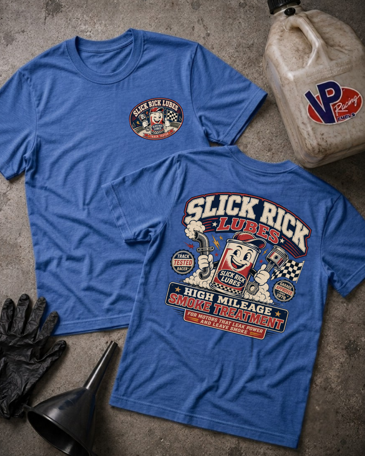 Two blue graphic T-shirts laid flat on a textured gray surface, with a black glove, funnel, and a dirty plastic jugs beside them. The front shirt shows a small circular 'Slick Rick Lube' logo on the left chest, and the back shirt shows a large 'Slick Rick Lube' grease-station style graphic with cartoon characters and text including 'High Mileage Smoke Treatment.