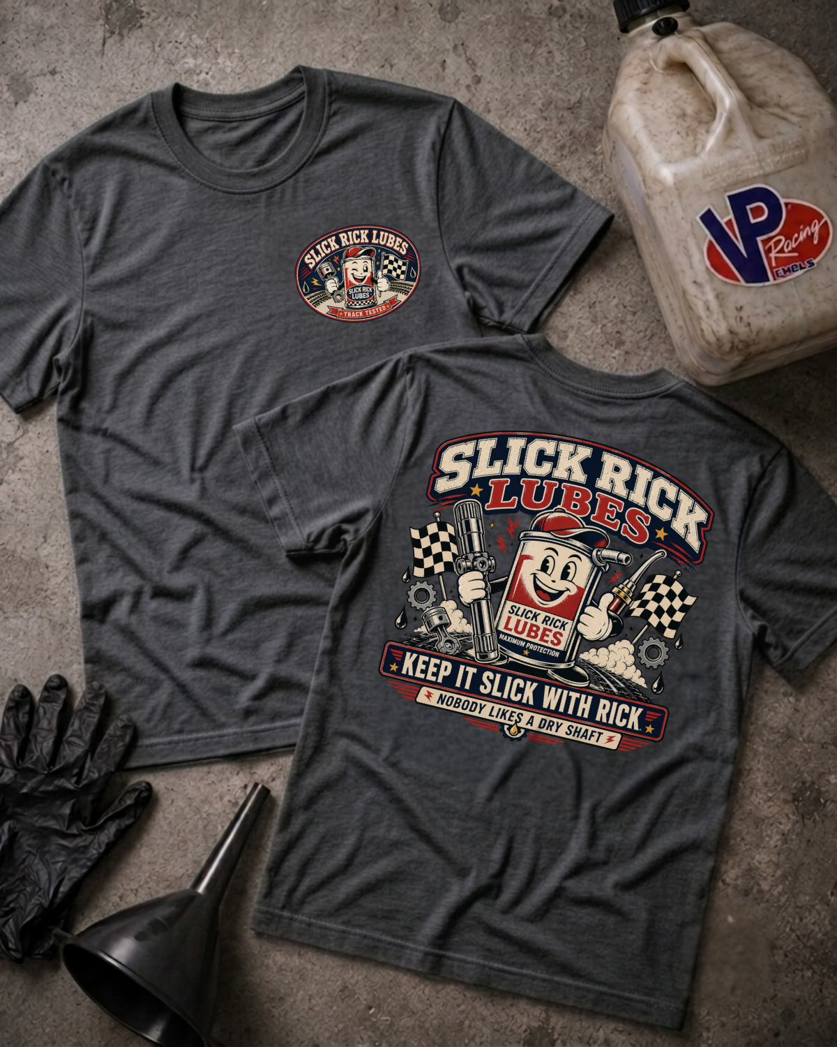 Two dark charcoal gray graphic T-shirts laid flat on a textured concrete surface, showing both front and back designs for Slick Rick Lubes. A beige VP Racing fuel jug, black gloves, and a black funnel are placed around them as styled props.