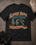 A black graphic t-shirt laid flat on a textured gray surface, featuring a vintage-style design with the words 'Slippery Nipple' and 'Grease Fittings' above illustrations of metal fittings and a grease fitting, with a banner reading 'Grab 'em and lube, she will love it too.' Black gloves and a funnel are placed around the shirt, with a beige plastic jug in the top right corner.