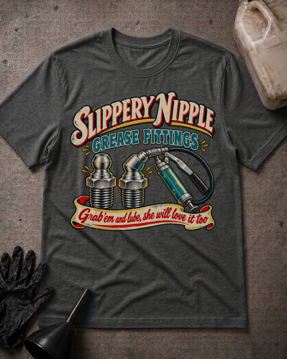 A dark gray graphic t-shirt laid flat on a textured gray surface, featuring a vintage-style grease fitting illustration with the text 'Slippery Nipple Grease Fittings' and 'Grab em and lube, she will love it too.' A black glove, funnel, and plastic jug are placed around the shirt as styled props.