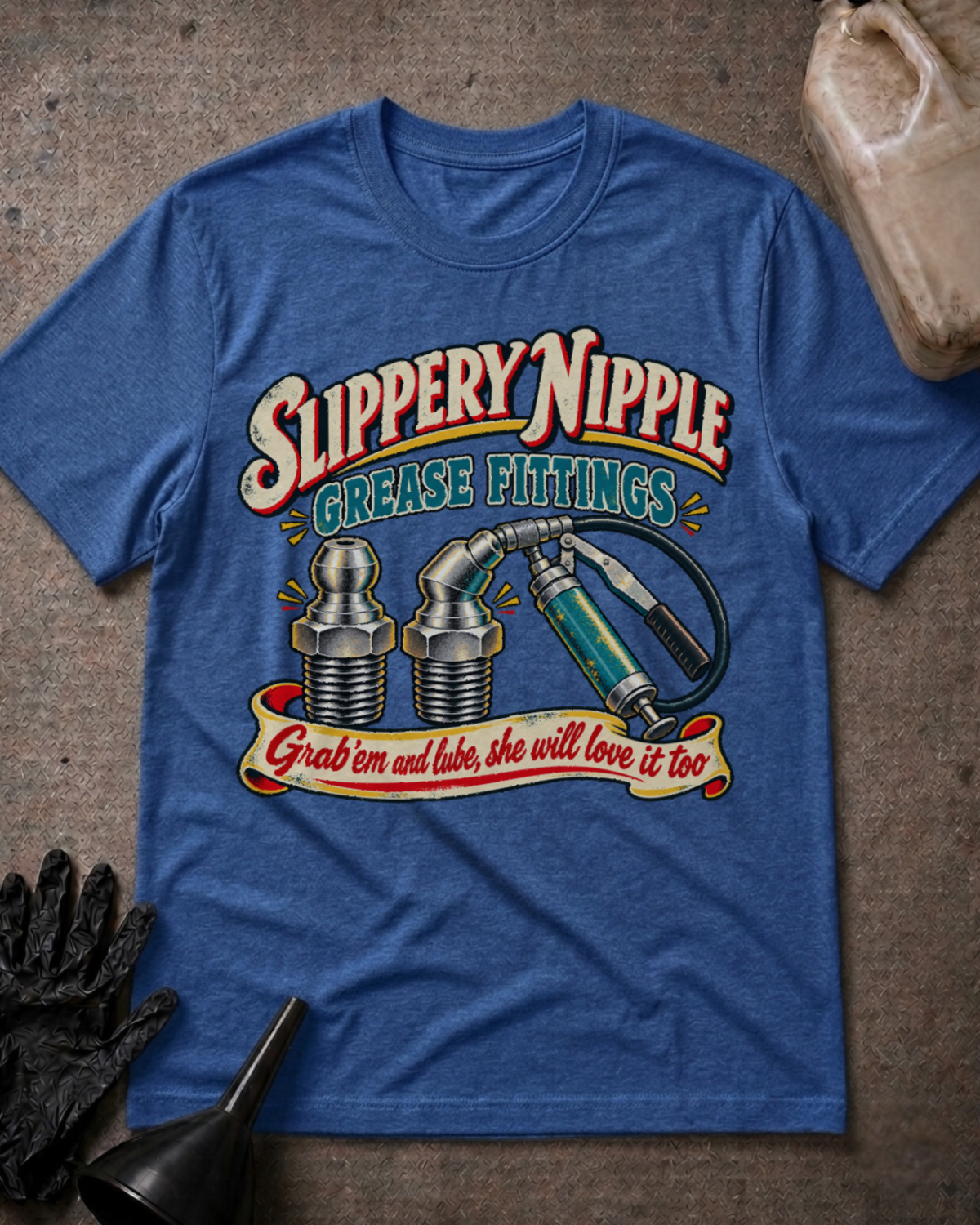 A heather blue graphic t-shirt laid flat on a textured brown surface, styled with a black work glove, a funnel, and a plastic bottle. The shirt features a retro-style graphic with the text 'Slippery Nipple Grease Fittings' and 'Grab em and lube, she will love it too' above illustrations of grease fittings and a grease pump.