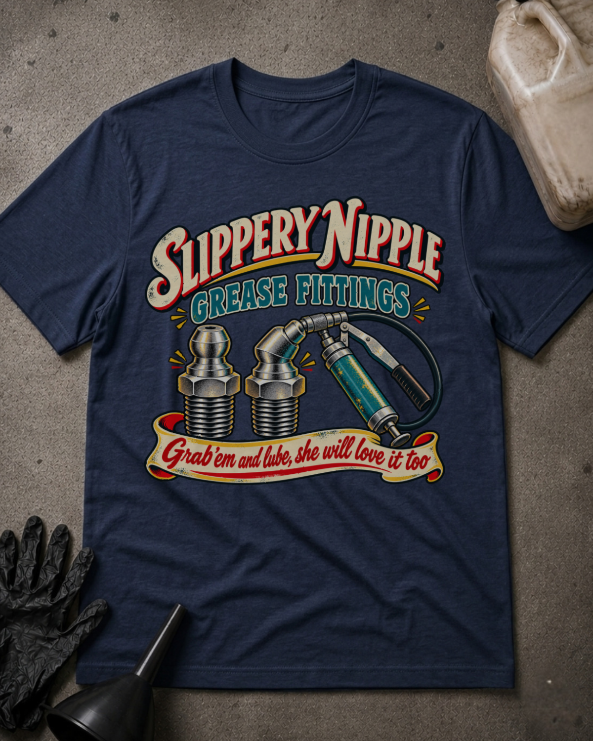 A navy blue graphic t-shirt laid flat on a textured gray surface, surrounded by automotive-themed props including a black glove, funnel, and plastic jug. The shirt features a retro-style graphic with the text 'Slippery Nipple Grease Fittings' and 'Grab 'em and lube, she will love it too' above illustrations of a grease fitting, a syringe, and a wrench.