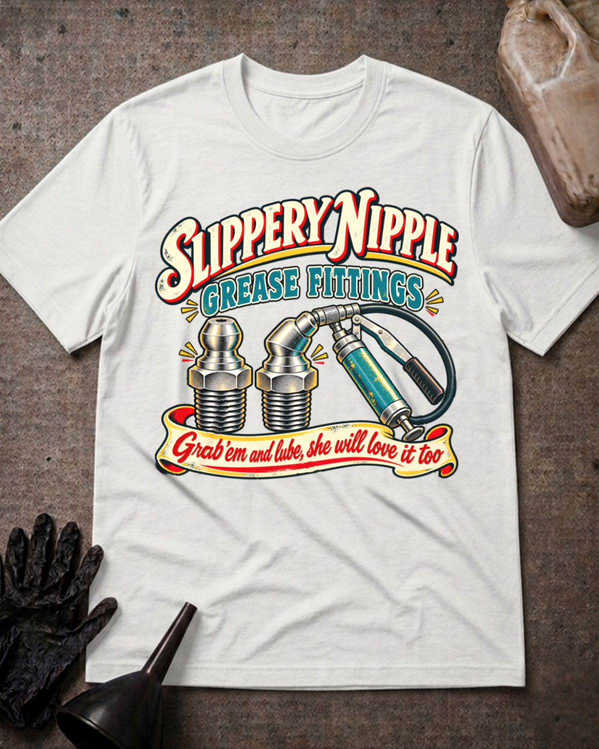 A white graphic t-shirt laid flat on a textured gray surface, featuring a vintage-style oil change illustration with the text 'Slippery Nipple Grease Fittings' and 'Grab em and lube, she will love it too.' A black glove, funnel, and wood block are styled around the shirt.