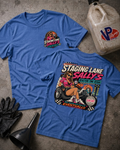 Two blue graphic T-shirts laid flat on a textured gray surface, with a vintage gas can, black gloves, and a funnel nearby. The front shirt shows a large retro race graphic reading 'Staging Lane Sally's' with a pin-up car scene, and the back shirt has a smaller matching chest graphic.
