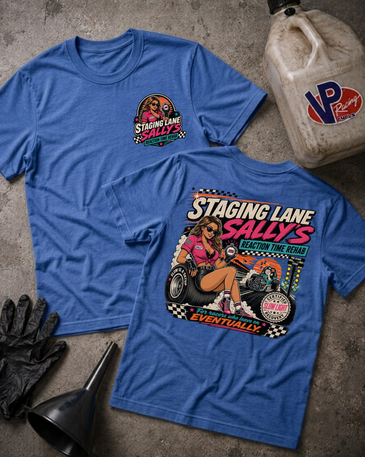 Two blue graphic T-shirts laid flat on a textured gray surface, with a vintage gas can, black gloves, and a funnel nearby. The front shirt shows a large retro race graphic reading 'Staging Lane Sally's' with a pin-up car scene, and the back shirt has a smaller matching chest graphic.