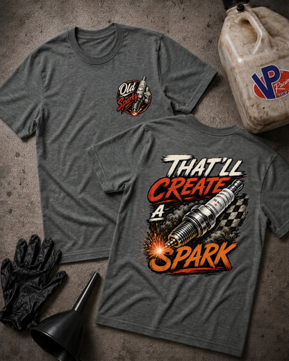 Two heather gray graphic T-shirts laid flat on a concrete surface, with automotive-themed props including black gloves, a black funnel, and a dirty plastic jug. The front shirt shows a small chest graphic reading Old Sparky, and the back shirt has a large spark-themed graphic with the text 'THAT'LL CREATE A SPARK.