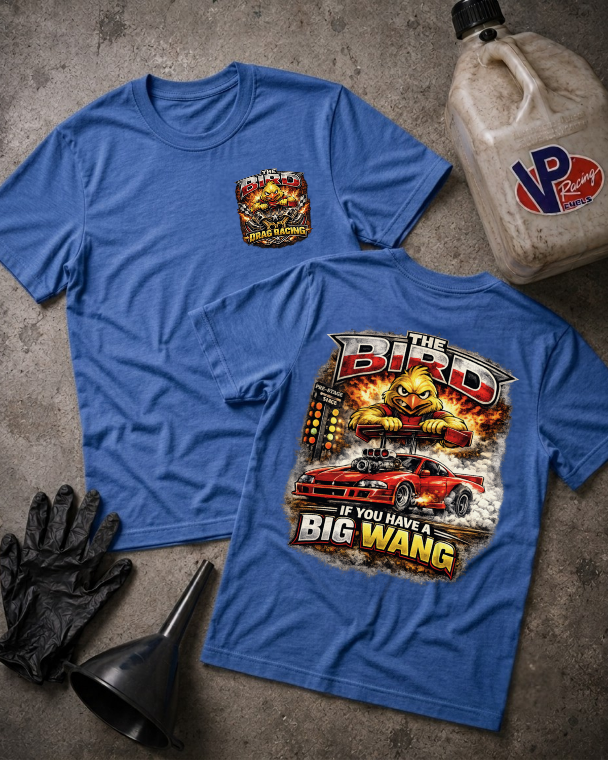 Two blue graphic T-shirts laid flat on a gray concrete-style surface, with racing-themed artwork and text including 'The Bird' and 'If You Have A Big Wang.' A VP Racing jug, black gloves, and a black funnel are placed around them as props.