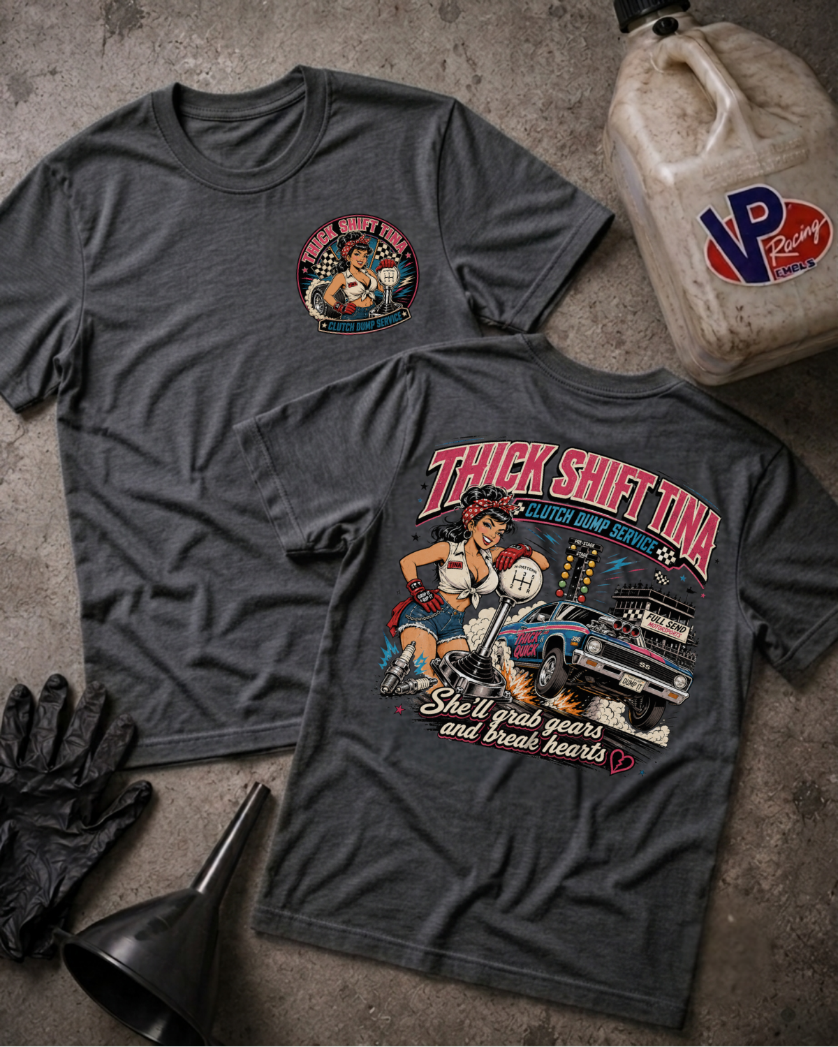 Two dark heather gray graphic T-shirts laid flat on a textured concrete surface, with a vintage oil-change illustration on the back of one shirt and a circular chest graphic on the other. A VP Racing fuel container, black gloves, and a black funnel are arranged around them as styled props.