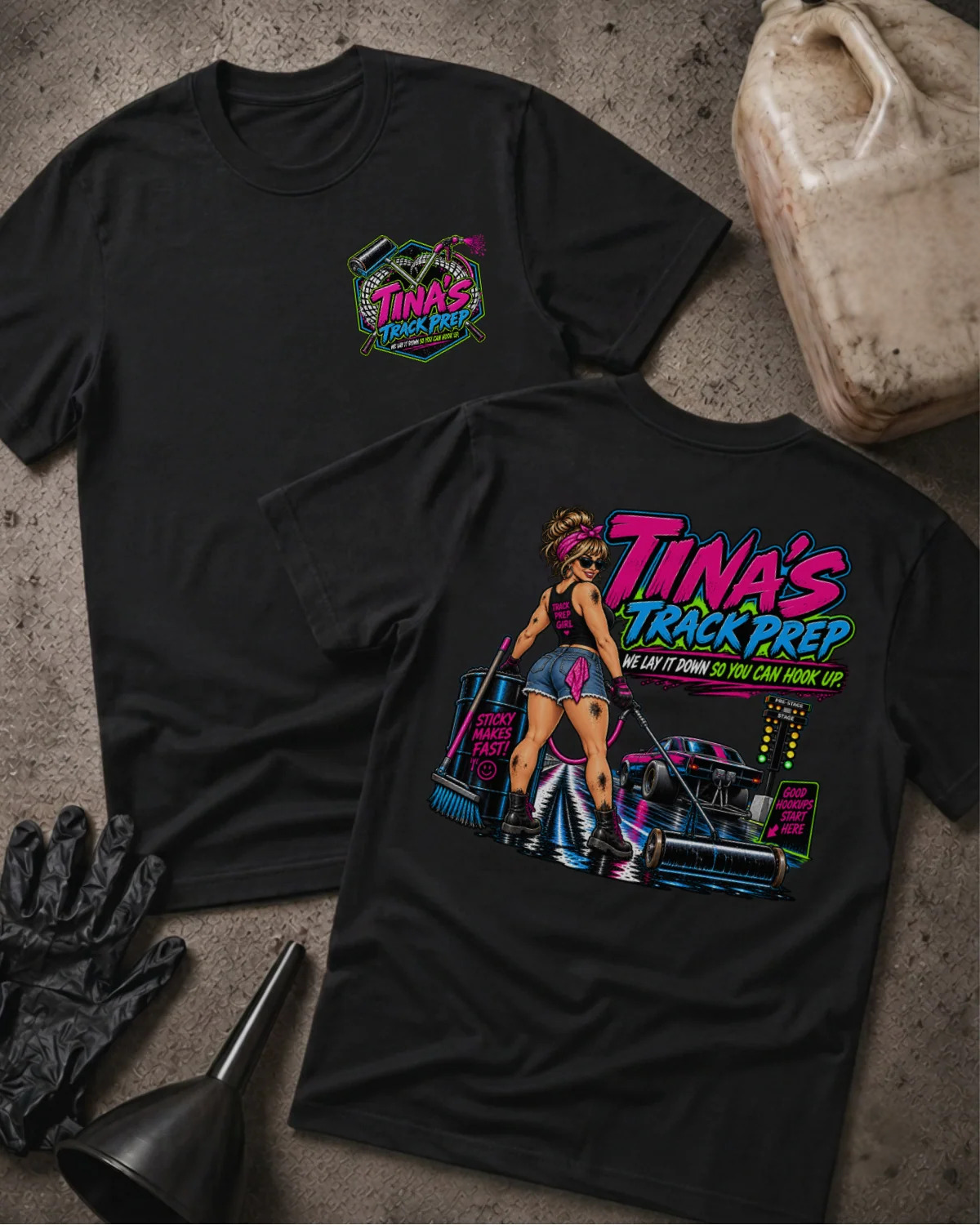 Two black graphic T-shirts laid flat on a textured concrete surface, with a gas can, black gloves, and a funnel nearby. The front shirt shows a small multicolor chest graphic reading 'Tina's Track Prep,' and the back shirt shows a large colorful graphic with the same text plus an illustrated woman and a race car.