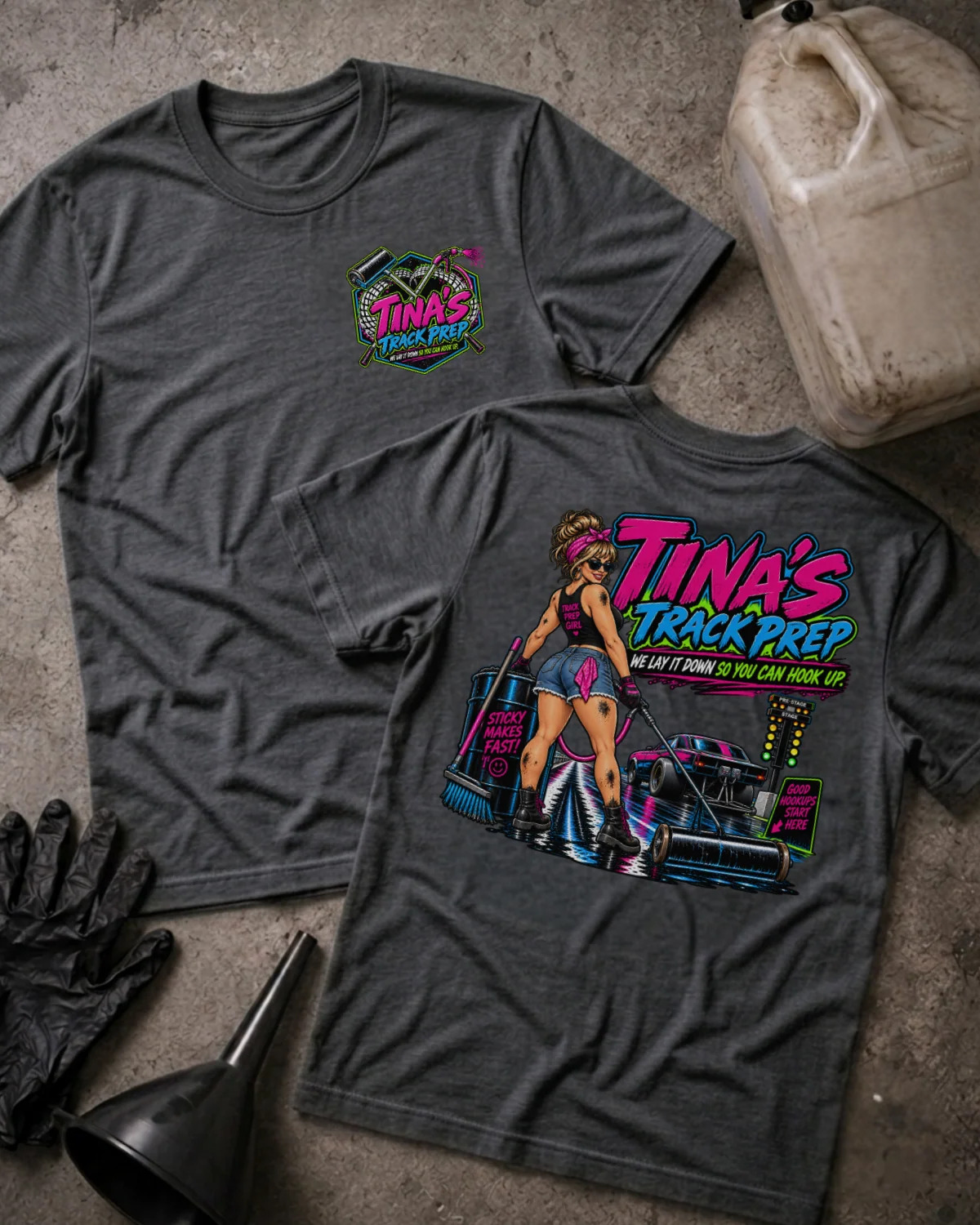 Two dark grey heather graphic T-shirts laid flat on a textured concrete surface, with a gas can, black gloves, and a funnel nearby. The front shirt shows a small multicolor chest graphic reading 'Tina's Track Prep,' and the back shirt shows a large colorful graphic with the same text plus an illustrated woman and a race car.